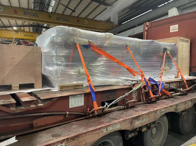 An oversized industrial mold wrapped in protective plastic being secured with orange ratchet straps and chains on a flatbed trailer for export logistics.